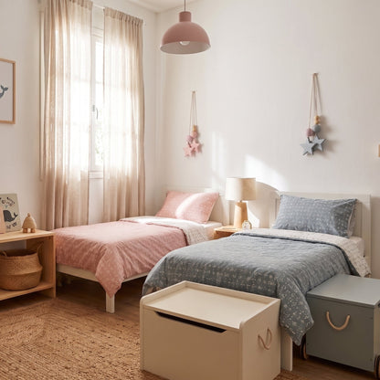 A styled children's bedroom featuring two single beds with pink and blue-gray bedding, a wooden shelf with toys, a bean bag, and a storage box, all in a warm neutral color palette.