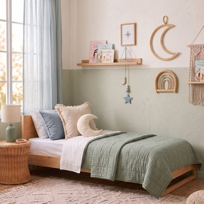 A cozy children's bedroom featuring a single wooden bed dressed in sage green quilted bedding, white and light blue pillows, and a crescent moon pillow. The room has a textured two-tone wall in cream and pale sage green, decorated with a wooden shelf, a macrame wall hanging, and a wicker side table with a lamp.