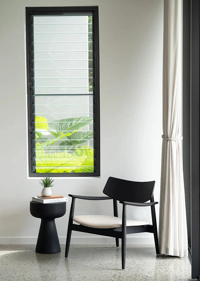 Modern black chair with white cushion next to a window with blinds and a white curtain.