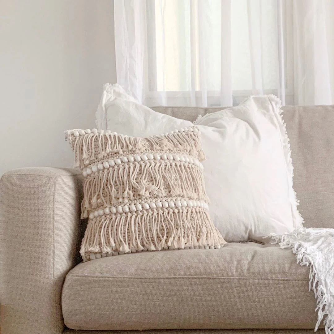 Beige sofa with textured pillow and white blanket in a room with light filtering through sheer curtains.