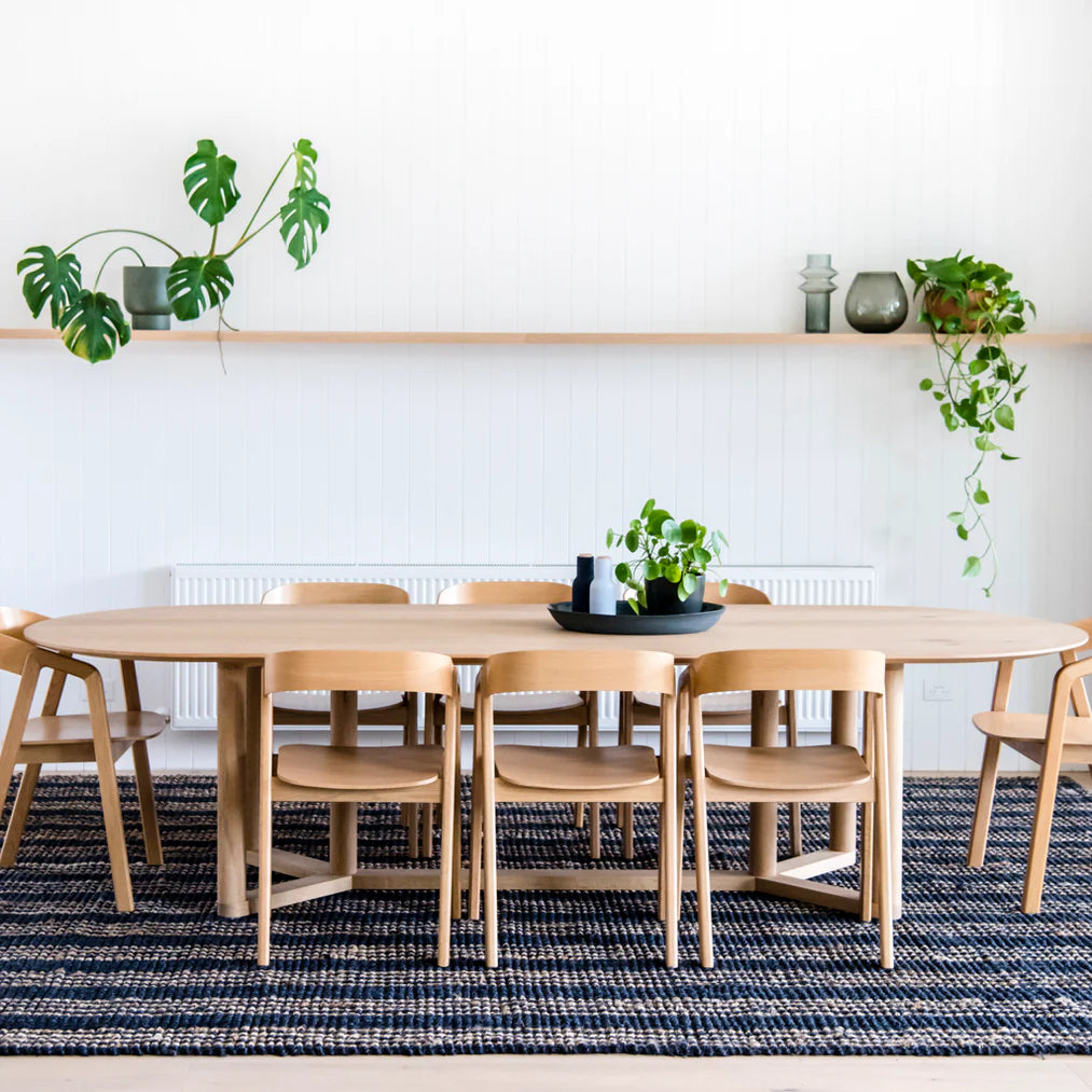 Dining area with wooden table and chairs, plants, and a shelf against a white wall.
