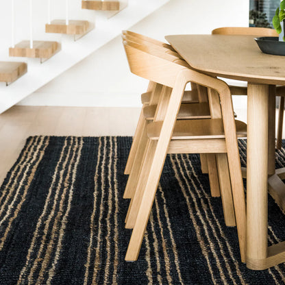 Wooden dining table and chairs on a black and gold striped rug with a white wall and staircase in the background.