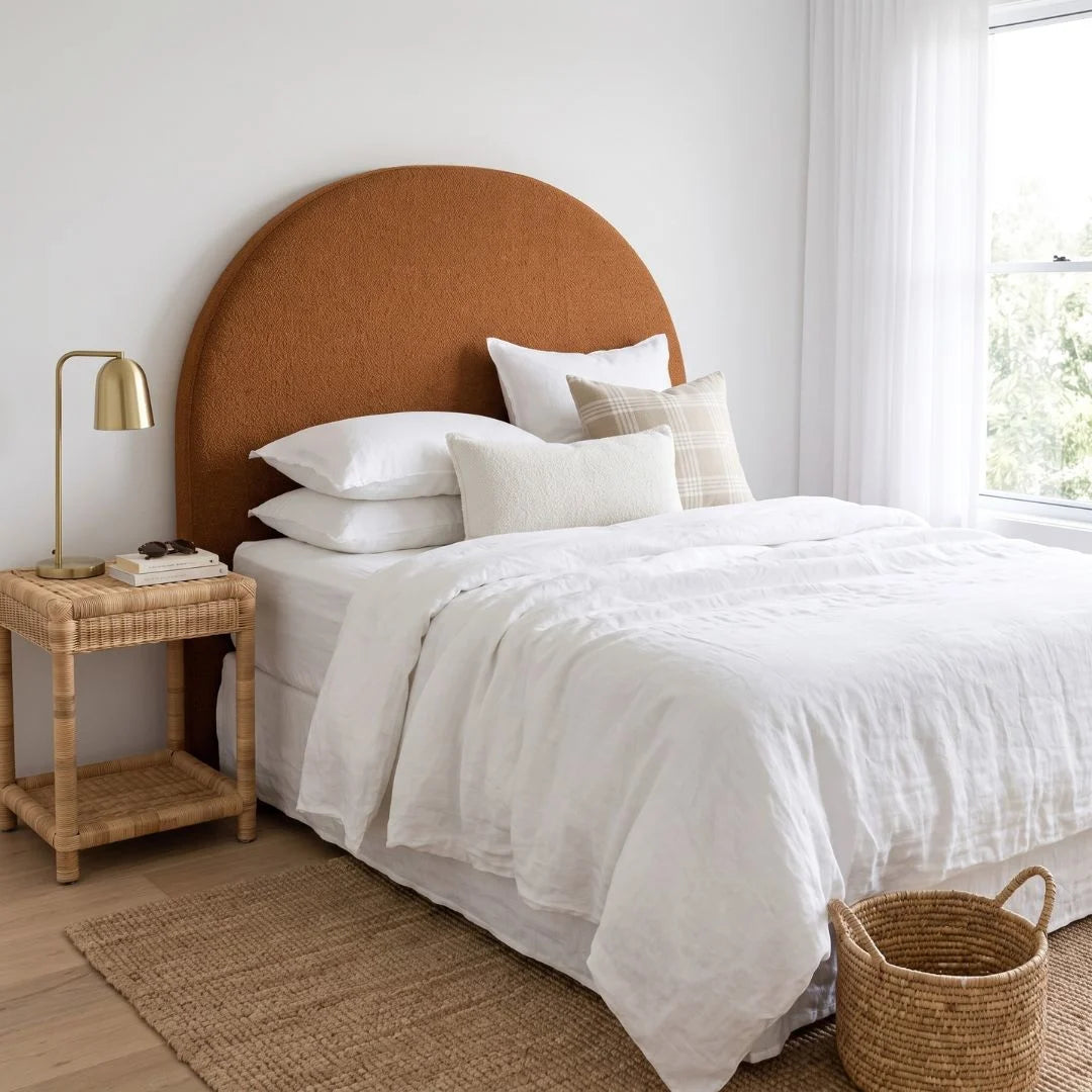 A styled bedroom scene featuring a large, semi-circular upholstered headboard in a warm rust-brown color, dressed in white bedding with decorative pillows. A matching woven rattan nightstand sits on the left, and a natural jute rug and woven basket are on the floor.