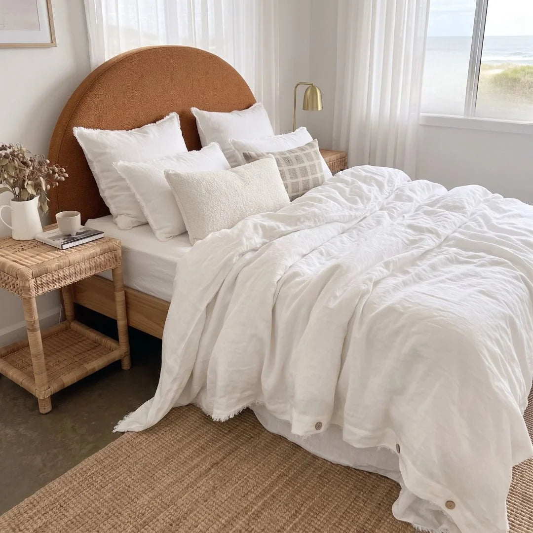 A neatly made bed in a bright bedroom setting, featuring white textured bedding, multiple white pillows, a round natural wood headboard, a rattan bedside table with a mug and dried flowers, sheer curtains, and a window looking out to the sea.