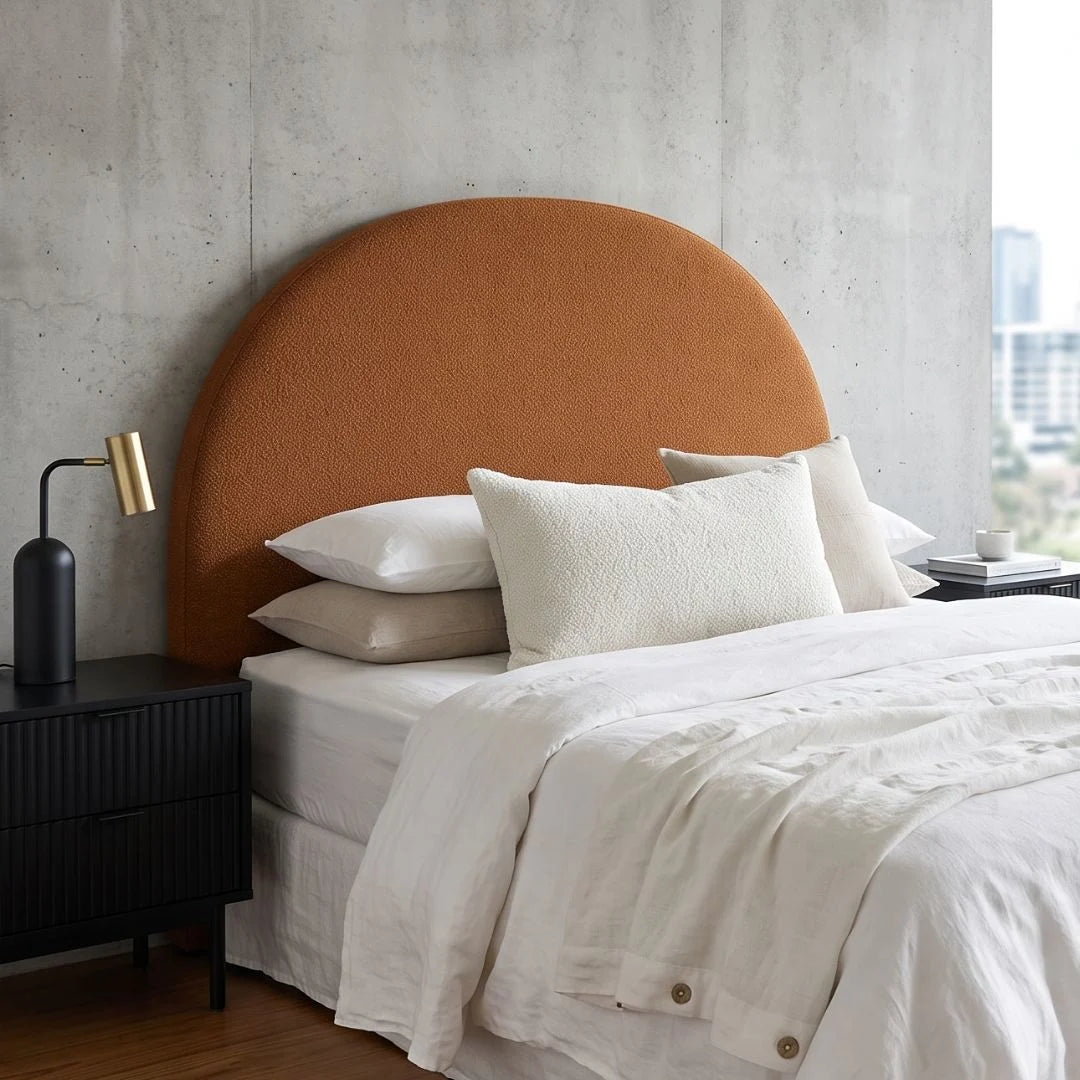 A modern bedroom scene featuring a large, semi-circular upholstered headboard in a warm terracotta orange fabric, styled with white and beige bedding, white pillows, a black bedside table with a gold lamp, and a concrete wall background.