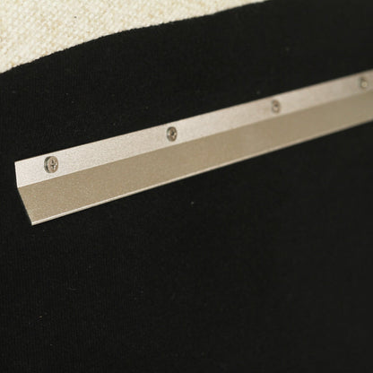 Close-up of a metallic silver metal track or bar with screws, mounted against a black textured background with a cream fabric corner visible at the top left.