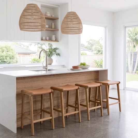 A bright modern kitchen island with a white countertop, light wood base, and four wooden bar stools with tan seats, set in a sunlit white kitchen with woven pendant lights.