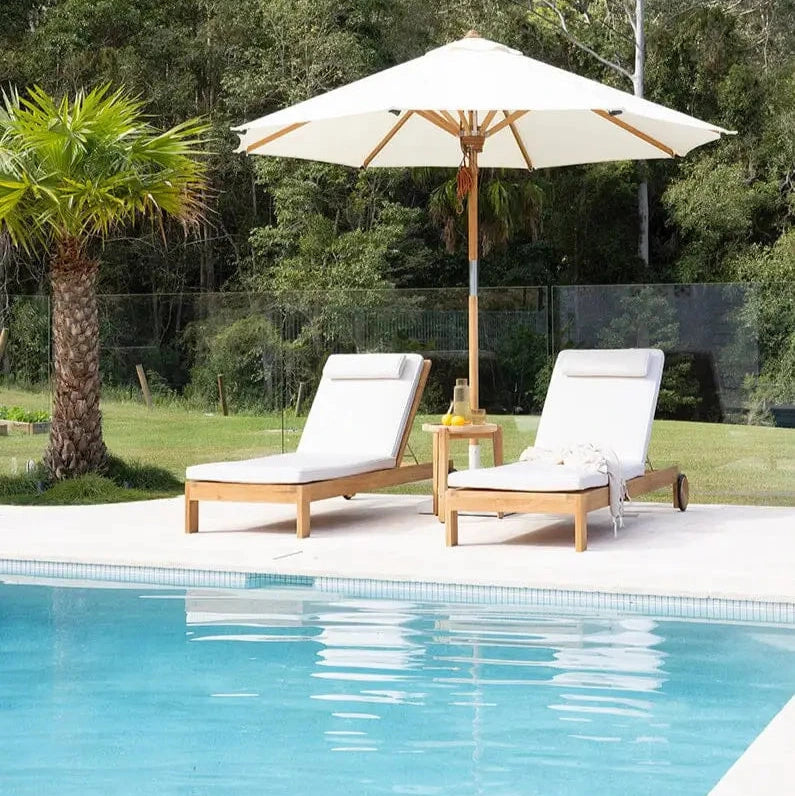 Two white lounge chairs with a beige umbrella by a poolside with greenery in the background.
