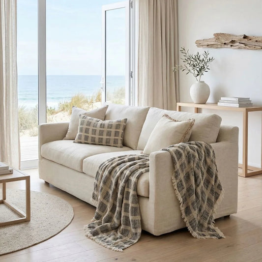 Beige sofa with plaid blanket in a living room with large windows overlooking a beach.