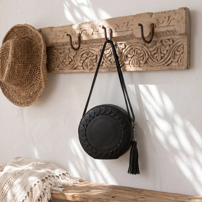 A black round shoulder bag with a tassel hangs from a carved wooden wall rack with hooks. A woven straw hat is also hanging on the left, with a textured cream blanket draped over a wooden bench in the foreground against a white wall with leaf shadows.