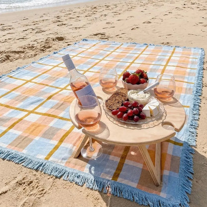 Picnic setup on a beach with a checkered blanket, table, wine, and food.