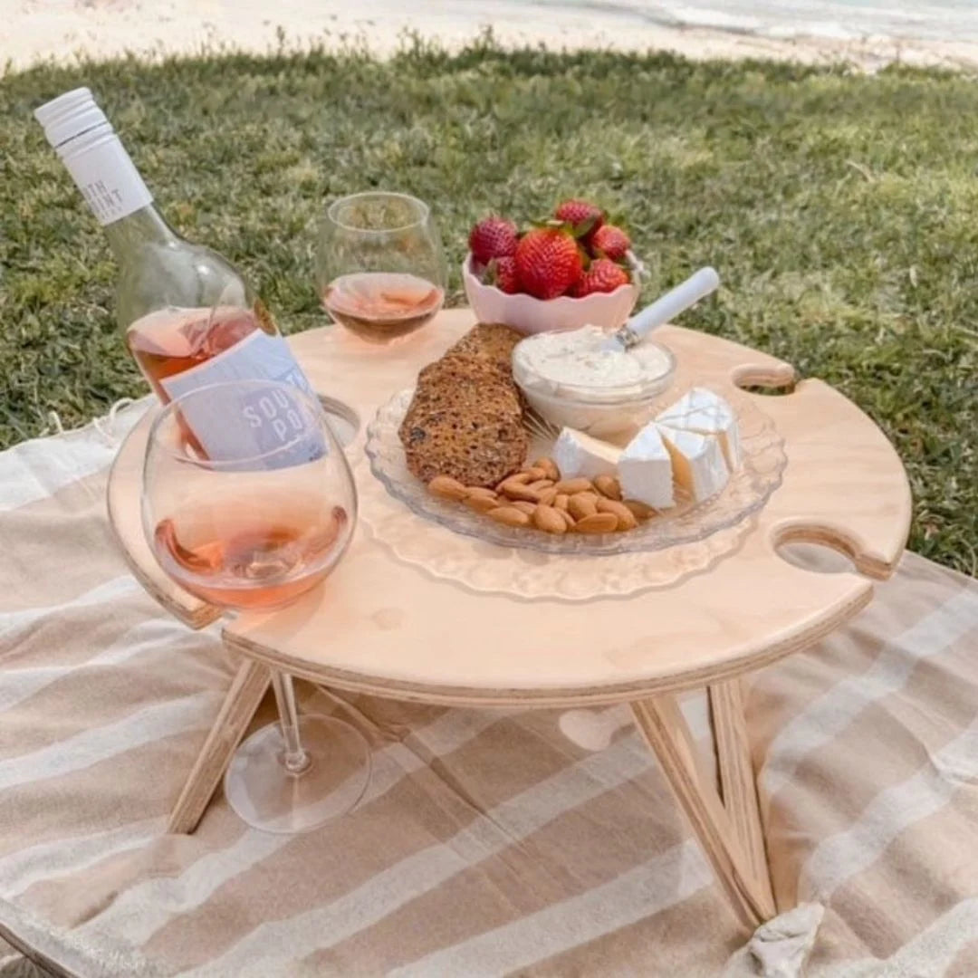 Picnic setup with a bottle of wine, glasses, fruits, and bread on a wooden tray outdoors.