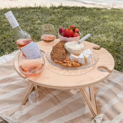 Picnic setup with a bottle of wine, glasses, fruits, and bread on a wooden tray outdoors.
