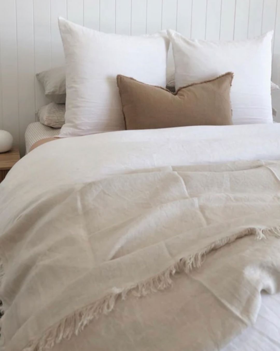 Neatly made bed with white and beige bedding against a white wooden panel wall.