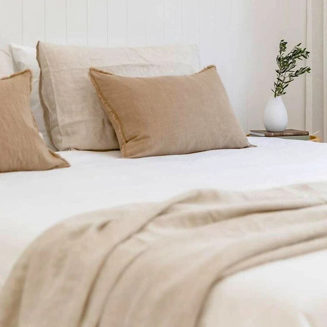 Neatly made bed with beige pillows and blanket, white nightstand with plant in the background.