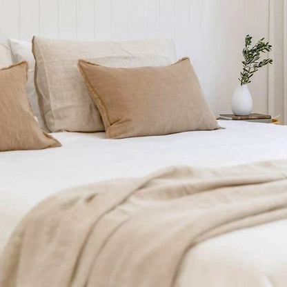 Neatly made bed with beige pillows and blanket, white nightstand with plant in the background.