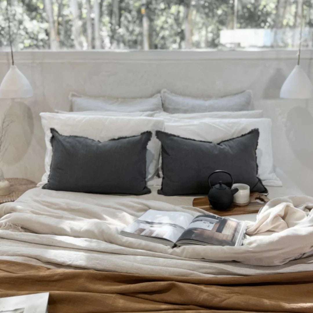 Bed with gray and white pillows, a book, and a teapot on a wooden nightstand.