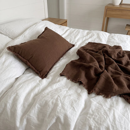 A brown textured pillow and matching throw blanket with frayed edges styled on a white bed in a bright bedroom setting.