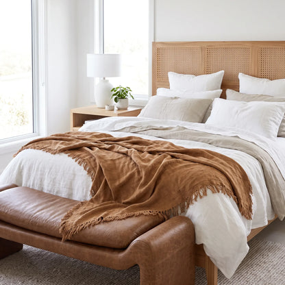 A styled bedroom scene with a bed dressed in white and beige linens, a rust-brown fringed throw blanket draped across the foot, a light wood headboard, and a brown leather bench at the foot of the bed. A white nightstand with a lamp and small plant sits beside the window.