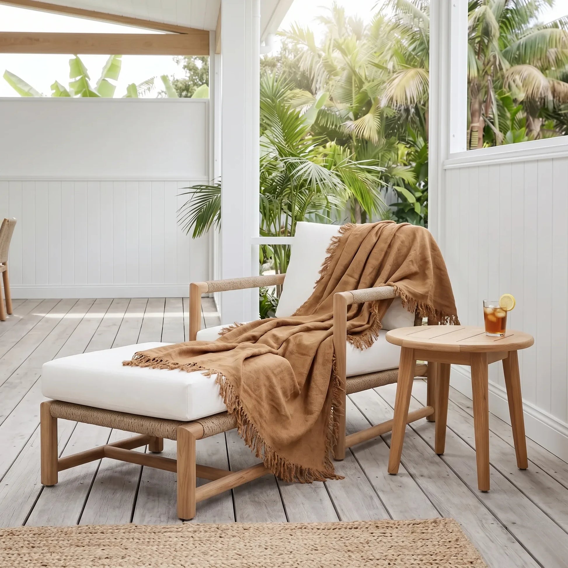 A light wood outdoor chaise lounge with white cushions and a tan fringed throw blanket, styled on a porch with a small matching side table and tropical greenery in the background.