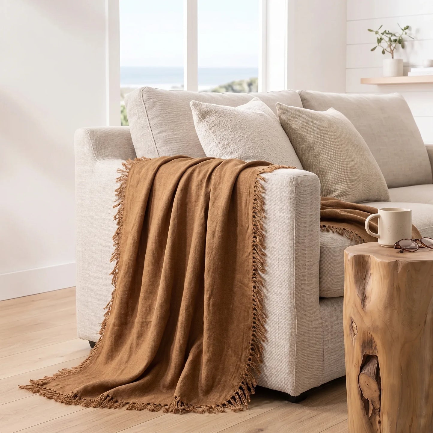 A warm camel brown throw blanket with fringe trim draped over a light beige fabric sofa in a bright, modern living room setting.