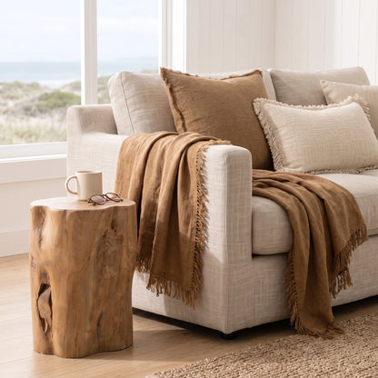 A styled living room scene featuring a light beige sofa with textured throw pillows and a warm brown fringed throw blanket, alongside a rustic natural wood side table holding a mug and sunglasses, with a window showing a beach view in the background.