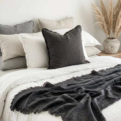 Bed with gray and white pillows and a dark gray throw blanket, with a vase of dried plants on a nightstand.