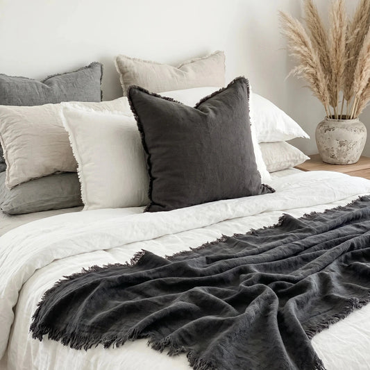 Bed with gray and white pillows and a dark gray throw blanket, with a vase of dried plants on a nightstand.