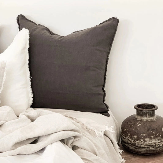 Charcoal Gray Ramie Linen Cushion  on a bed with white bedding and a rustic vase in the corner.