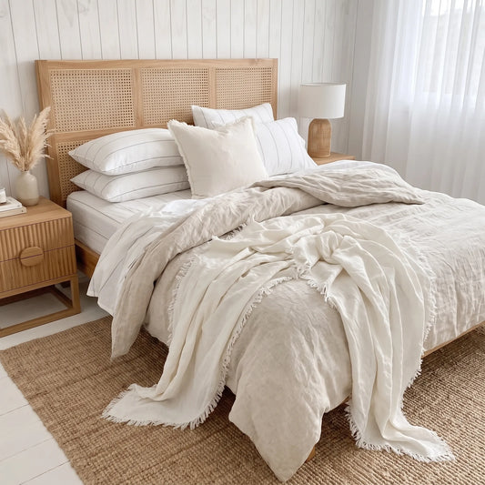 A neatly made bed in a bright, neutral-toned bedroom featuring a light wood headboard, white pillows, and cream-colored textured bedding with a fringed throw blanket draped over the foot of the bed.