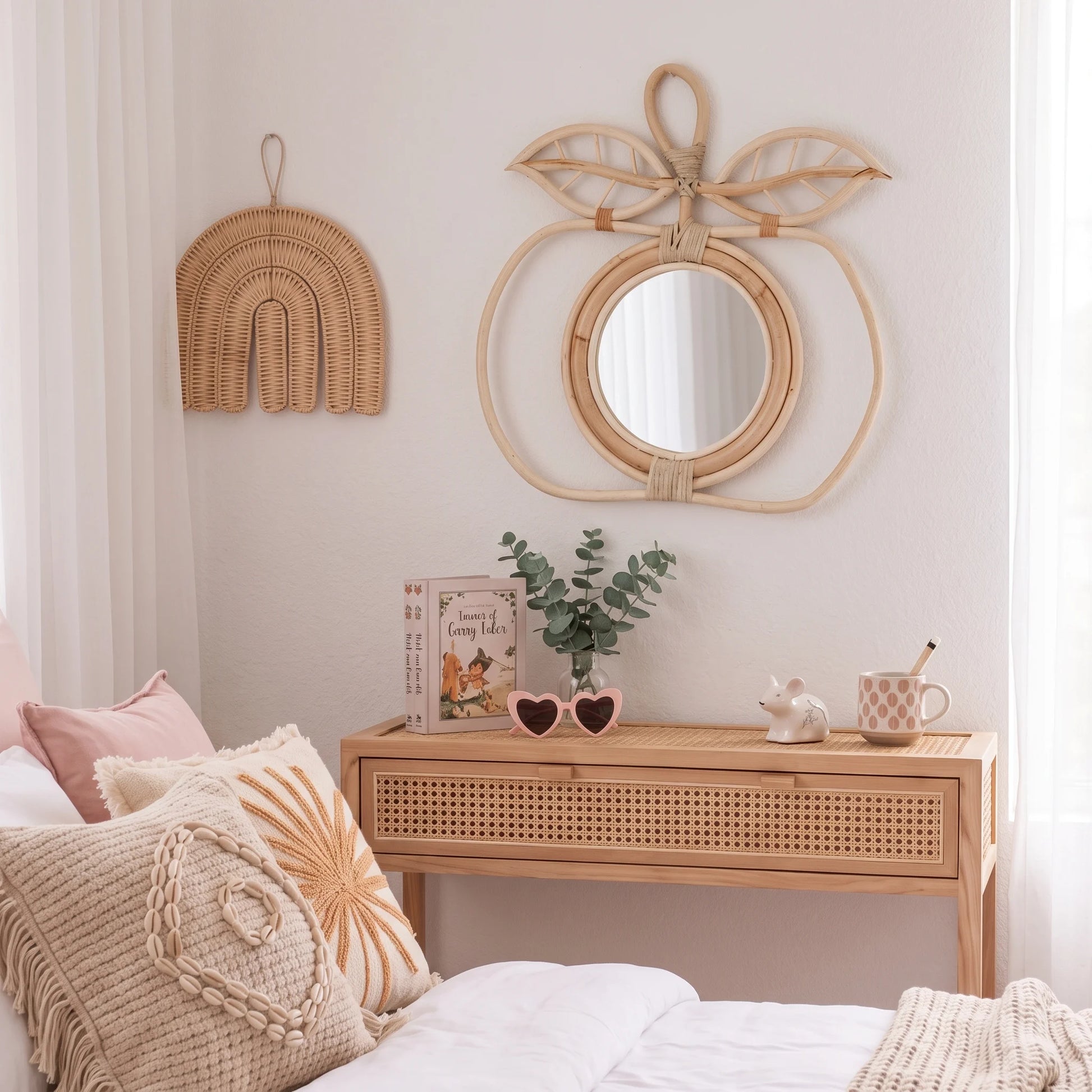 A styled bedroom corner featuring a light natural wood console table with a woven drawer front, a matching wicker wall mirror shaped like a peach, and a wicker rainbow wall hanging. The room has white walls, sheer curtains, and a bed with beige and pink pillows in the foreground.