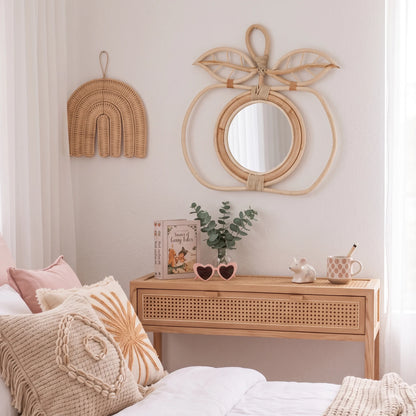 A styled bedroom corner featuring a light natural wood console table with a woven drawer front, a matching wicker wall mirror shaped like a peach, and a wicker rainbow wall hanging. The room has white walls, sheer curtains, and a bed with beige and pink pillows in the foreground.