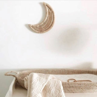 A woven crescent moon wall hanging in a natural straw color, mounted on a white wall above a matching woven changing tray with a textured cream blanket.