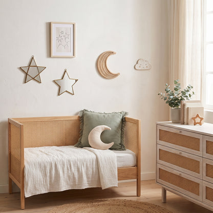 A styled children's bedroom corner featuring a light wood daybed with a woven cane frame, dressed in cream bedding and a sage green pillow with a crescent moon accent. A matching wooden dresser with woven drawer fronts stands beside a window with sheer curtains, decorated with a small plant and a star-shaped ornament.