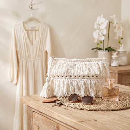 A cream-colored woven clutch bag with thick fringe tassels sits on a wooden table in a styled room setting, alongside sunglasses, a perfume bottle, and a woven placemat. A matching cream dress hangs on a hanger in the background.