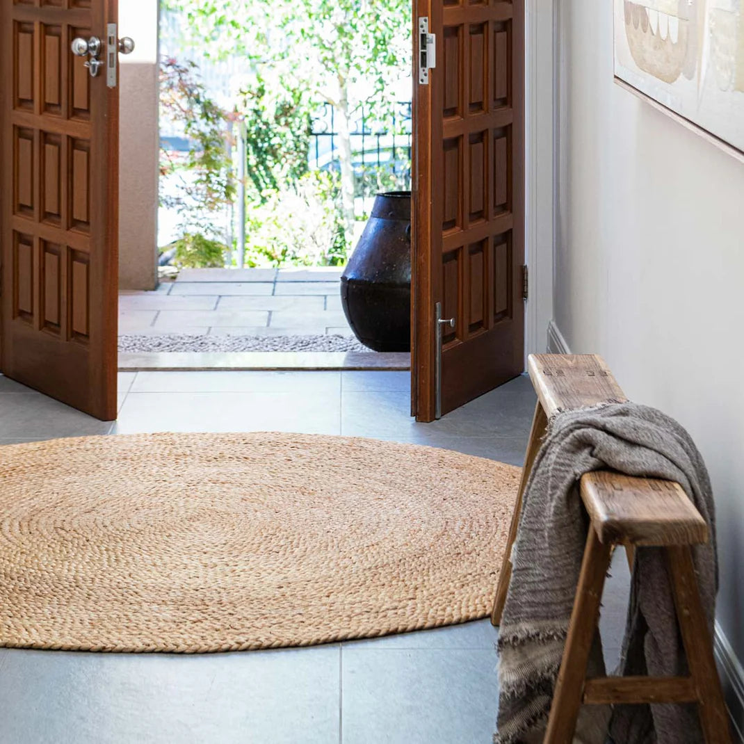 Entryway with a woven mat, wooden bench, and open doors leading to a garden.