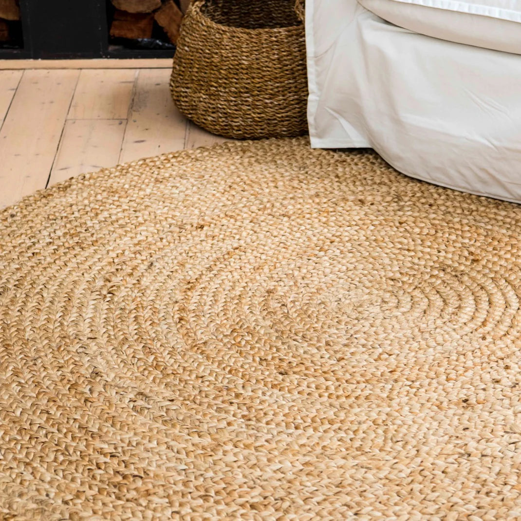 Round woven jute rug on a wooden floor with a basket and bed in the background.