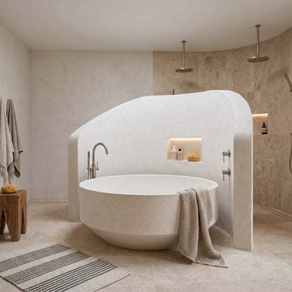A modern freestanding oval bathtub in a minimalist beige bathroom with a curved surround wall, built-in shelf, tiled walls, and a striped bath rug.