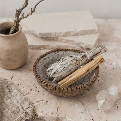 Smudge sticks in a ceramic bowl on a stone surface with a vase and crystals in the background.
