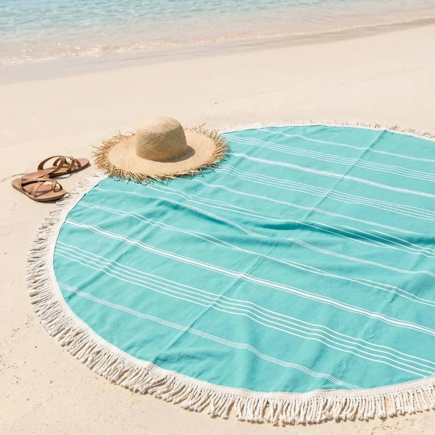 Round turquoise beach towel with white stripes on a sandy beach with a hat and sandals.