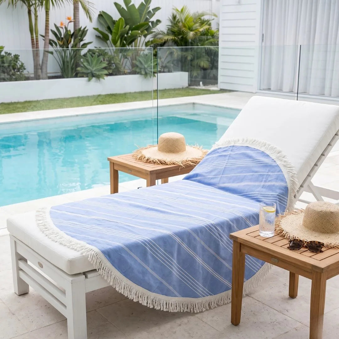 White lounge chair with a blue striped towel, straw hat, and sunglasses by a poolside.