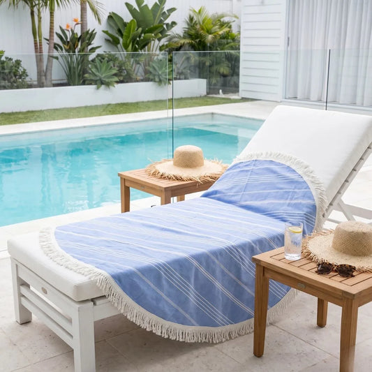 White lounge chair with a blue striped towel, straw hat, and sunglasses by a poolside.