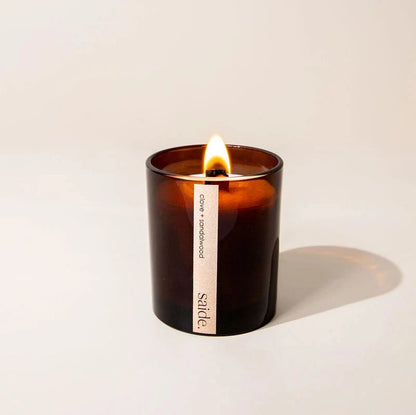 Candle in a brown glass jar with a visible brand label on a light beige background