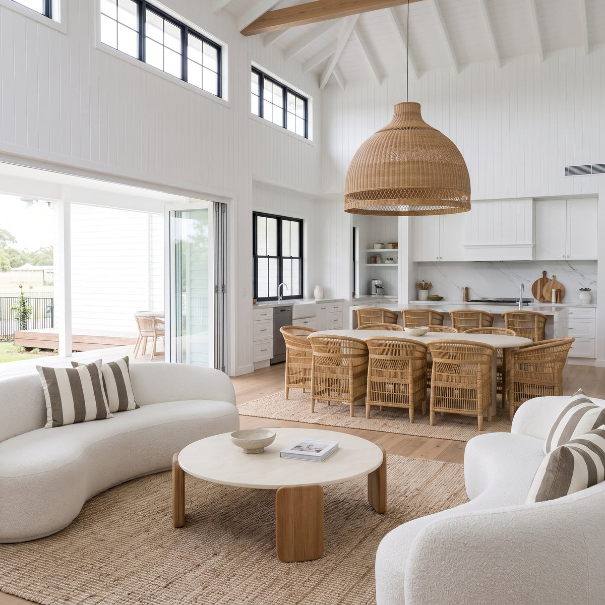 A bright, modern open-plan living space with a curved white sofa, round wooden coffee table, woven pendant light, dining set, and high white walls with tall windows.