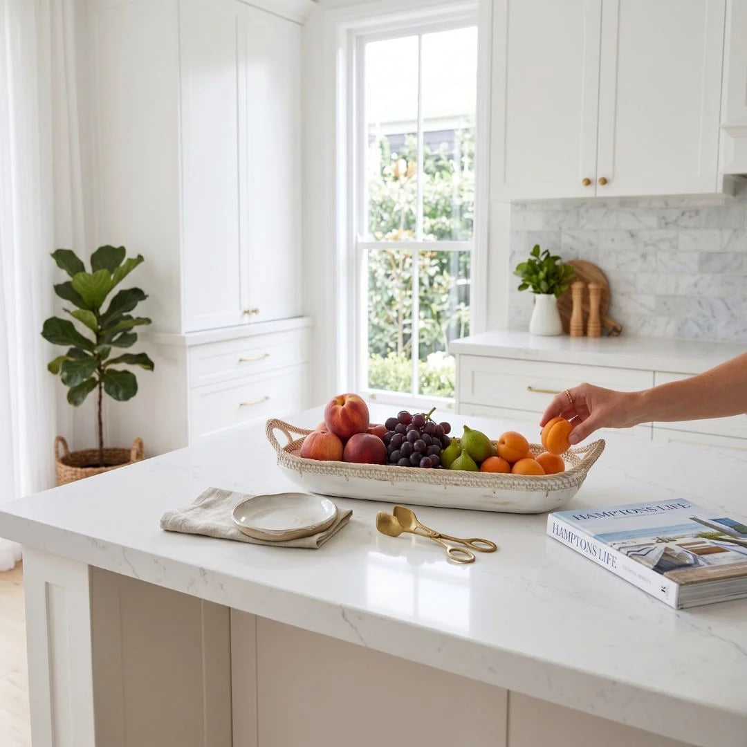 Person reaching for fruit on a kitchen counter with a bowl of fruit and a book.