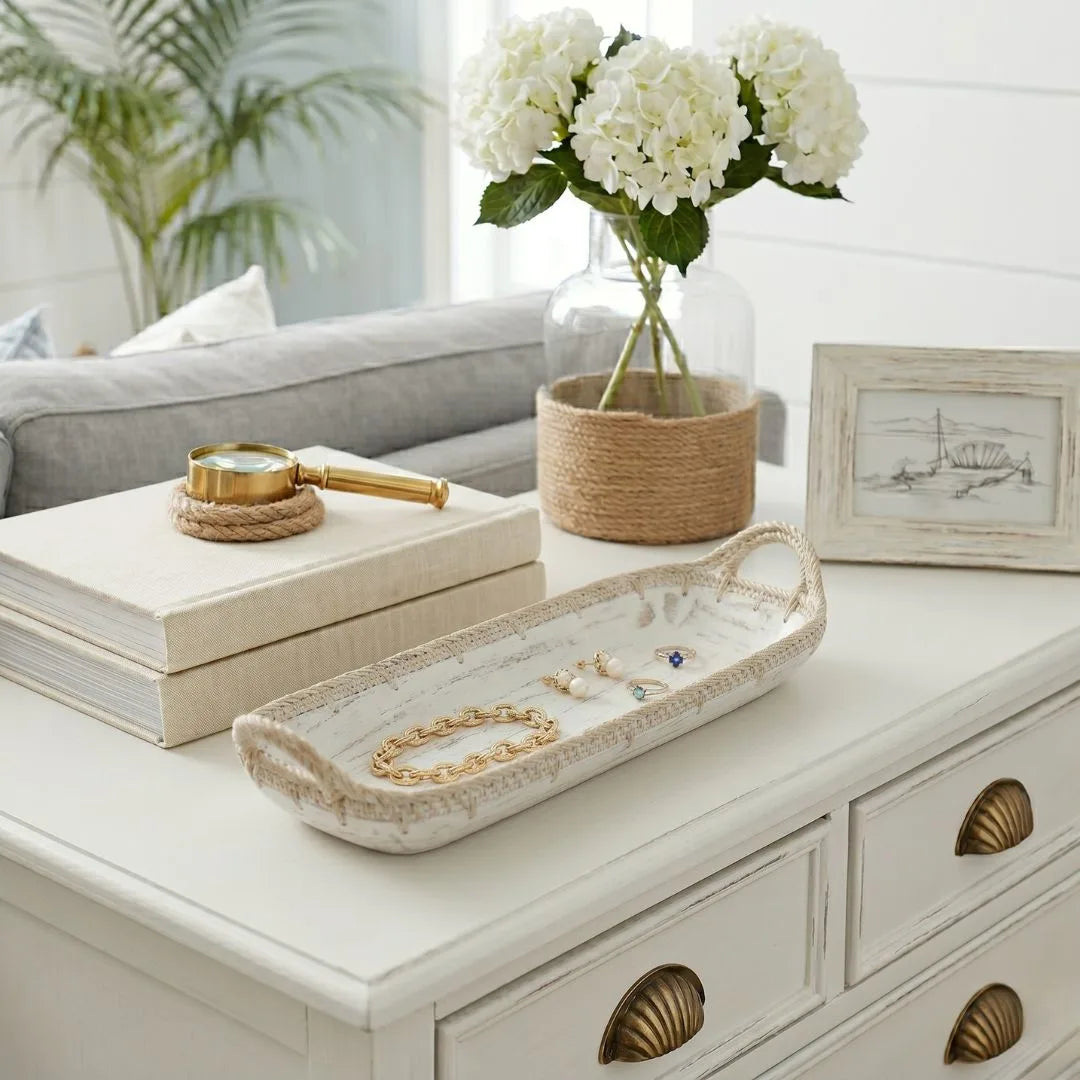 Decorative tray with jewelry on a dresser next to a vase of white flowers and books.