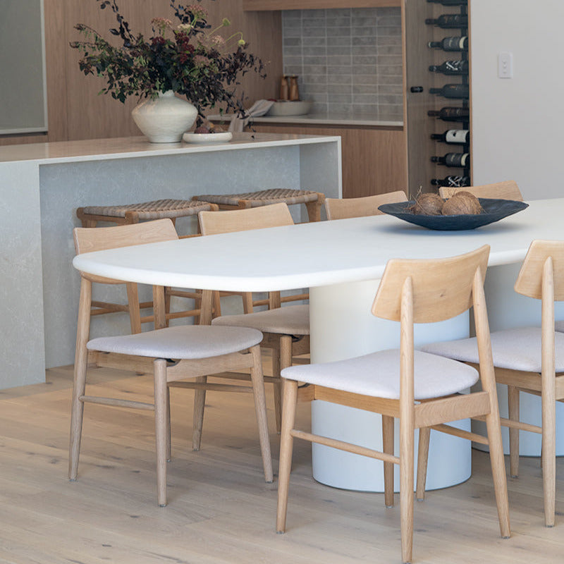 A modern dining room scene with a white oval dining table, light wood chairs with white upholstered seats, pendant lights, a kitchen island, and a wine rack in the background.
