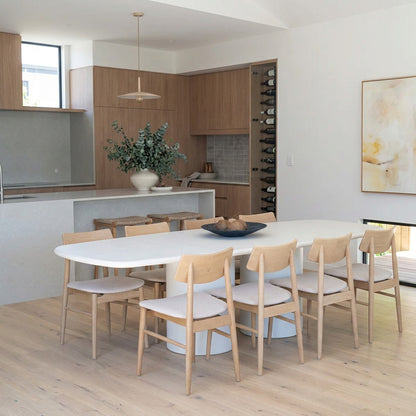 A modern dining room scene featuring a white oval dining table with a cylindrical base and six light natural wood chairs with white upholstered seats.