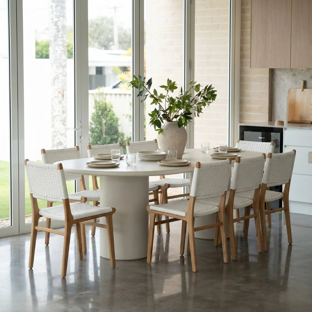 Dining room with a round white table and wooden chairs near large windows.