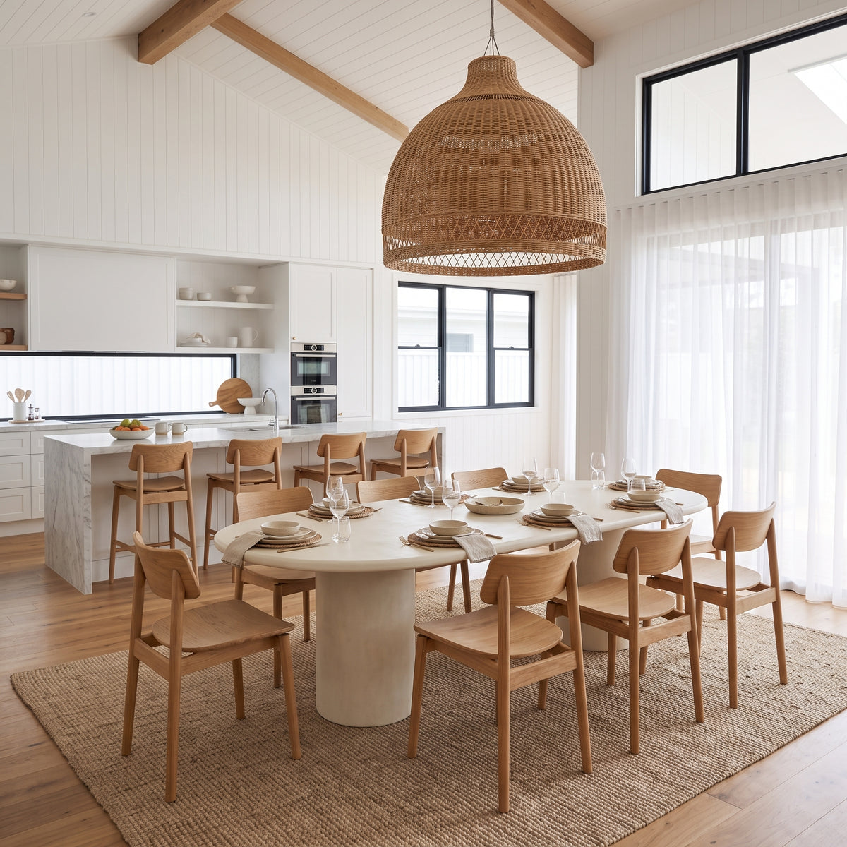 A modern dining room scene with a round white dining table surrounded by light wood chairs, under a large woven pendant light. A marble kitchen island and white cabinetry are visible in the background, along with large windows and sheer curtains.